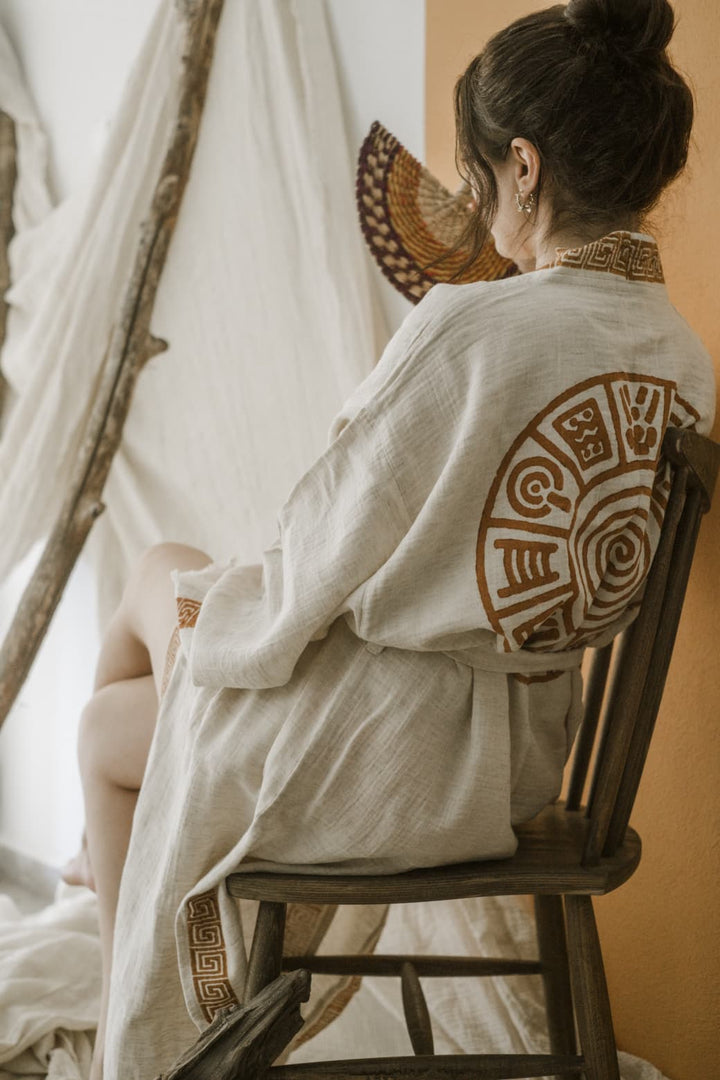 A woman with brown hair in a bun sits sideways on a wooden chair near white drapes and an orange wall, wearing the Triable Boho Kimono Robe - Poolside Beach Cover Up from turkanhome.com, featuring tribal hand block print.