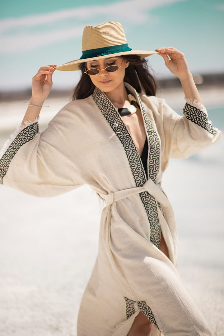 A woman models the Assos Lightweight Yoga Kimono by turkanhome.com with black trim, pairing it with a straw hat, sunglasses, and a chunky necklace outdoors against blue sky and white sand.