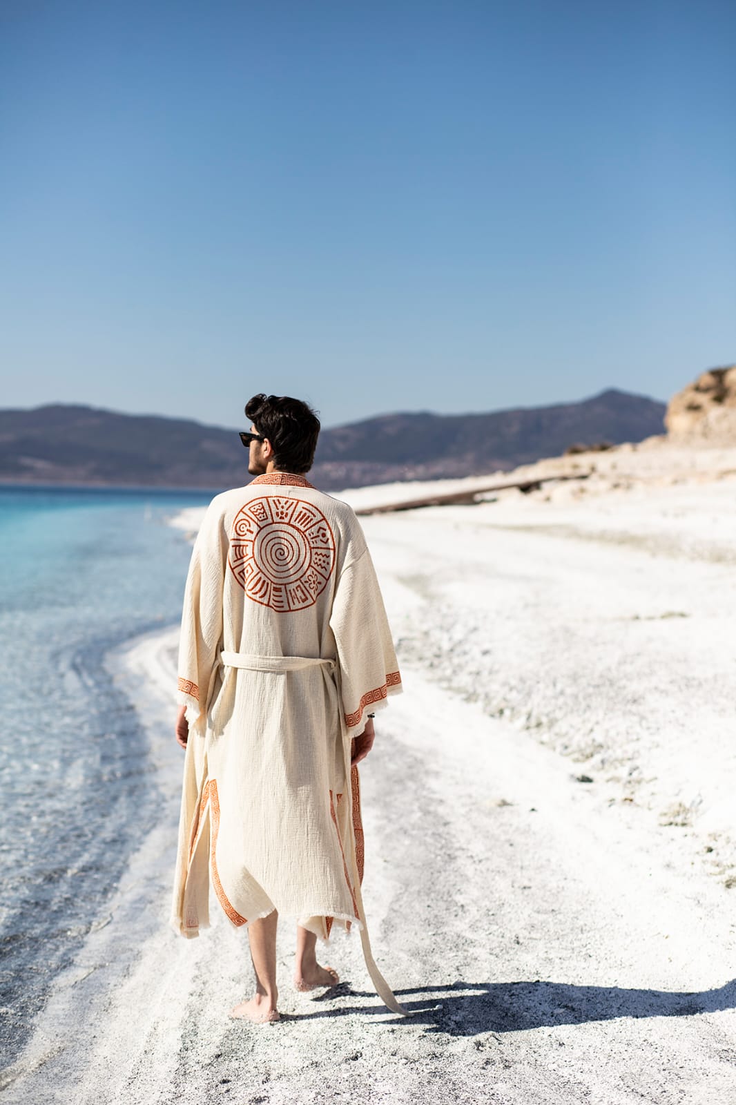 A man in the Triable Boho Kimono Robe by turkanhome.com, a cream cotton muslin cover-up with an orange circle on the back, walks barefoot along a sandy white beach near clear blue water under a bright sky, mountains distant.