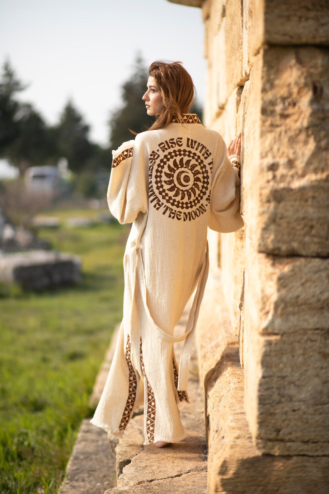 A woman wearing the Rise Kimono – a lightweight festival kimono from turkanhome.com with brown patterns and “RISE WITH THE MOON” on the back – stands outside by a stone wall, looking into the distance on a sunny day.