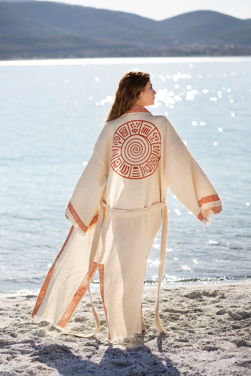 A woman wears the Triable Boho Kimono Robe by turkanhome.com, a long cream-colored beach cover-up with red tribal hand block print, as she stands on the sand facing the sea and hills under the sunlight.