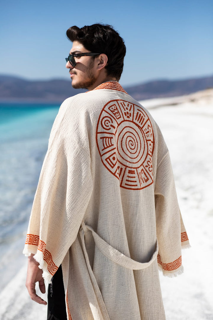 A man in the Triable Boho Kimono Robe by turkanhome.com, featuring orange geometric patterns, stands on a white sand beach near turquoise water. Mountains rise under a clear blue sky as he gazes into the distance, wearing sunglasses.