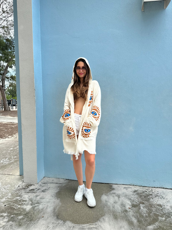 A woman with long hair and sunglasses wears the Mystic Eye Kimono Robe Short with Pockets and Hood by turkanhome.com over a white top, shorts, and sneakers, smiling outdoors against a light blue wall.