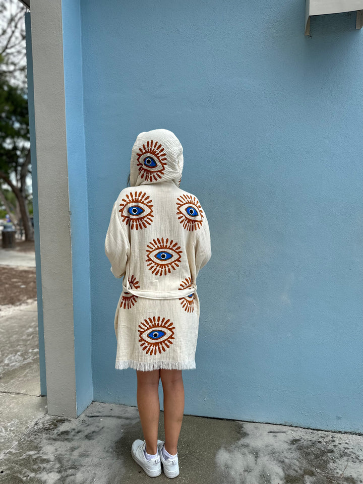 A person stands outdoors facing a light blue wall, wearing the Mystic Eye Kimono Robe Short with Pockets and Hood from turkanhome.com for an effortlessly eco-friendly look. The robe features large, colorful eye designs on cream cotton.