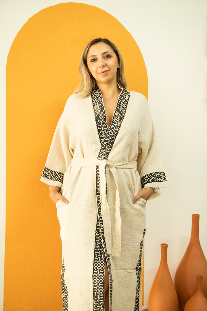 A woman wears the Assos - Lightweight Yoga Kimono by turkanhome.com, featuring beige fabric with patterned black trim, standing before an orange arch-shaped wall and three terracotta vases beside her.