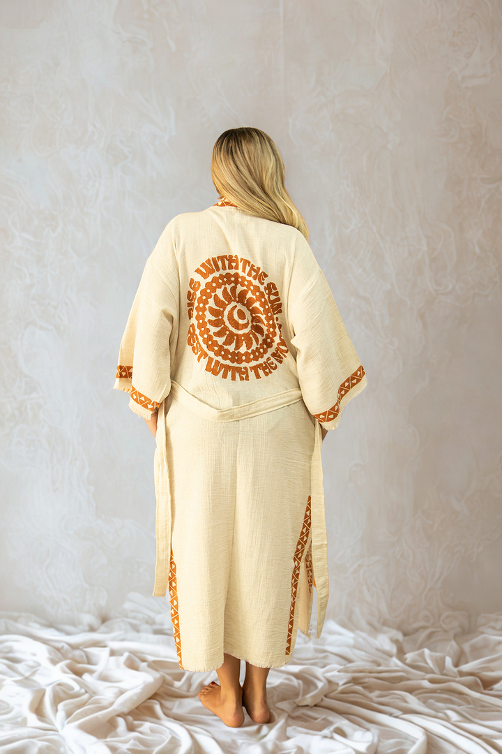 A woman with long blonde hair stands barefoot on white fabric, facing away. She wears the Rise Kimono, a lightweight festival kimono from turkanhome.com, featuring orange patterns and a bold sun design reading SHINE WITH THE SUN on the back.
