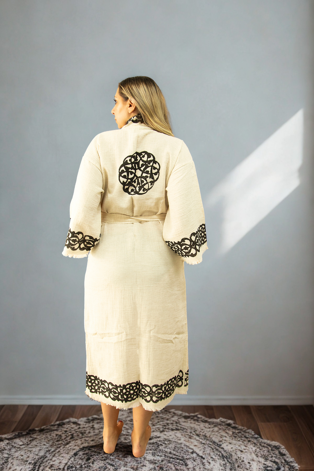 A barefoot woman stands on a round rug, facing away, wearing the Laodikya - Lightweight Yoga Kimono from turkanhome.com. The cream robe features black patterns on the sleeves, hem, and back, set against a sunlit neutral background.