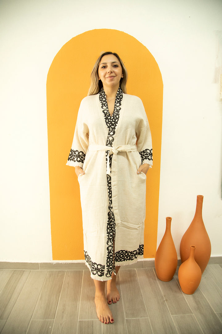 A woman stands barefoot indoors wearing the Laodikya Lightweight Yoga Kimono by turkanhome.com, cream-colored with black lace trim. She poses before an orange arch on a white wall, with three brown decorative vases nearby.