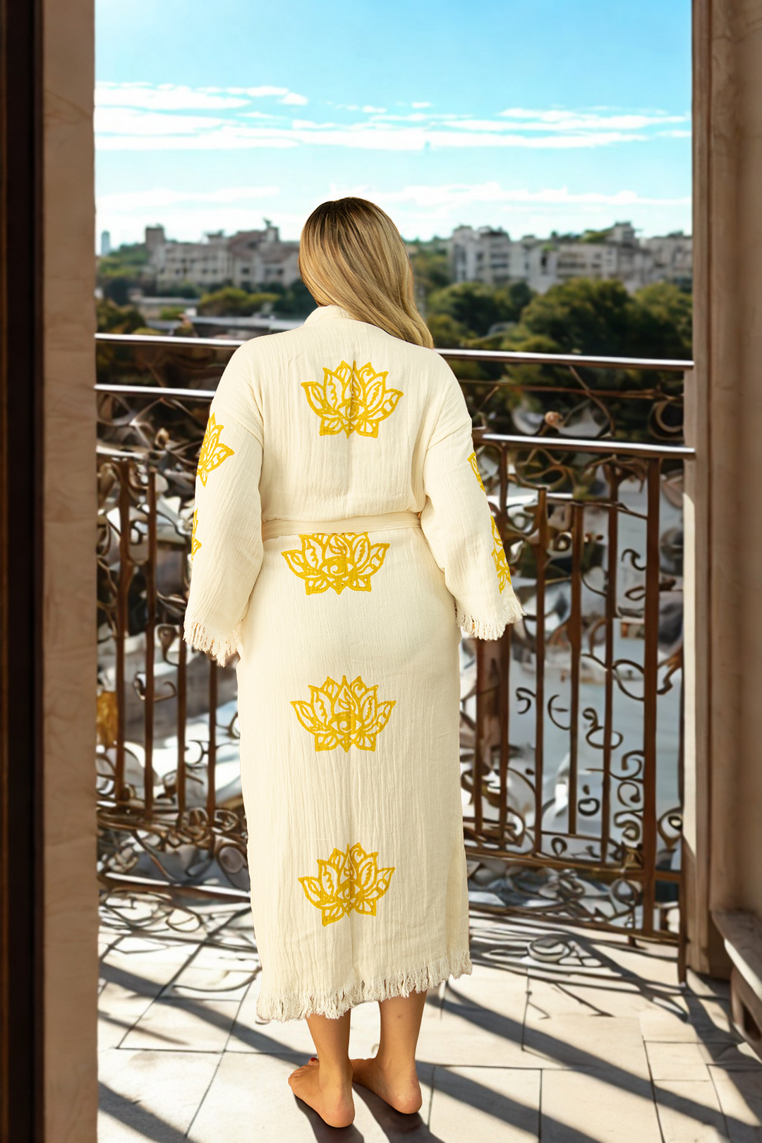 A barefoot blonde wearing the cream-colored Lotus Kimono Robe by turkanhome.com stands on a balcony, overlooking a sunny cityscape with distant trees and buildings.