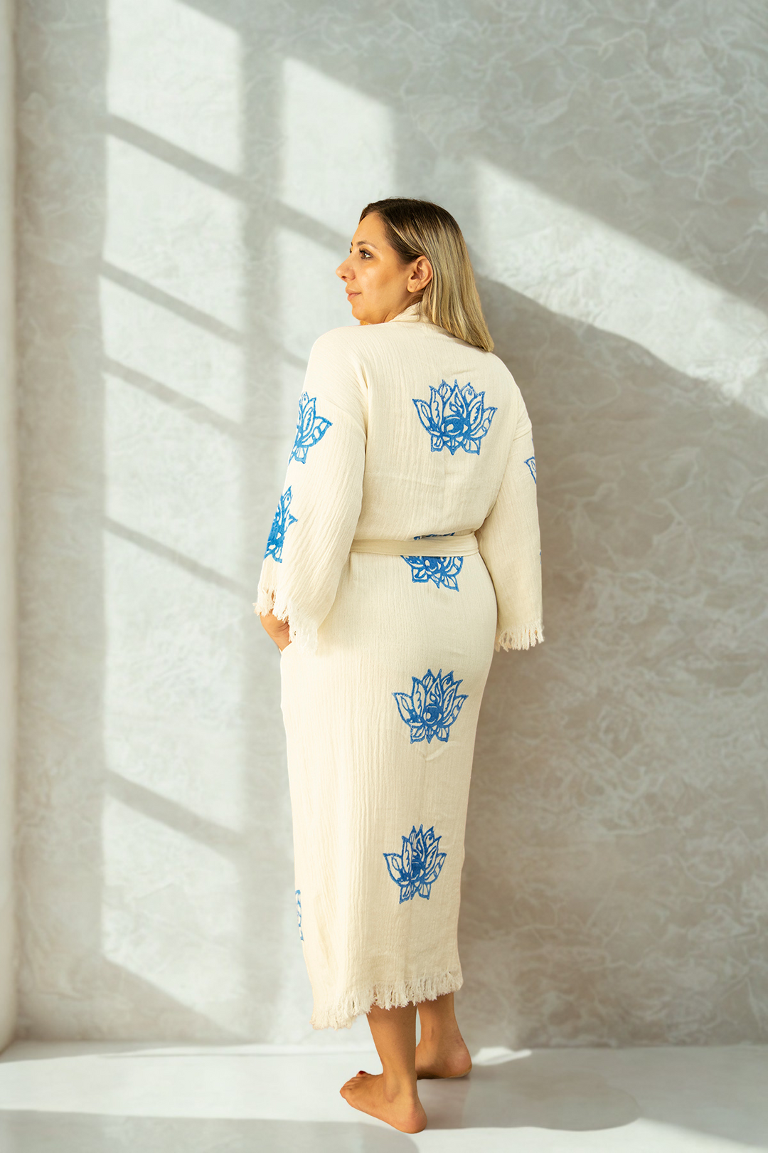 A woman stands barefoot on a white floor, back turned, wearing the eco-friendly Lotus Kimono Robe from turkanhome.com with blue lotus patterns. Sunlight casts soft shadows on the light-textured wall behind her.