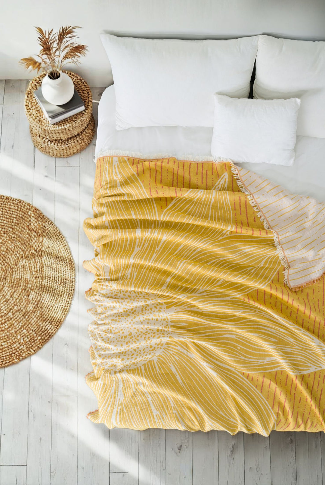 The Sunflower Bedspread in Yellow-Brick by turkanhome.com, made of 100% cotton with a large white jacquard floral pattern, decorates the cozy bed with white pillows, alongside a woven round rug and wicker side table on light wood flooring.