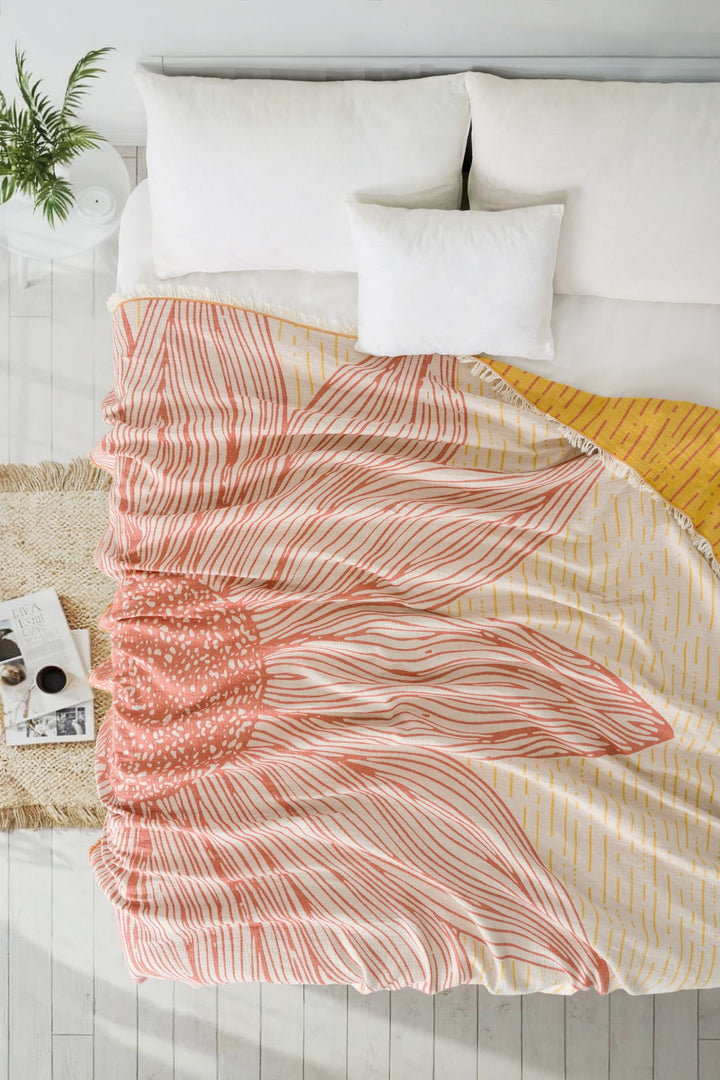 A neatly made bed features the Sunflower Bedspread in Yellow-Brick by turkanhome.com, 100% cotton with a pink and yellow jacquard pattern. White pillows, a woven rug, small table with books and photos, and a green potted plant complete the scene.