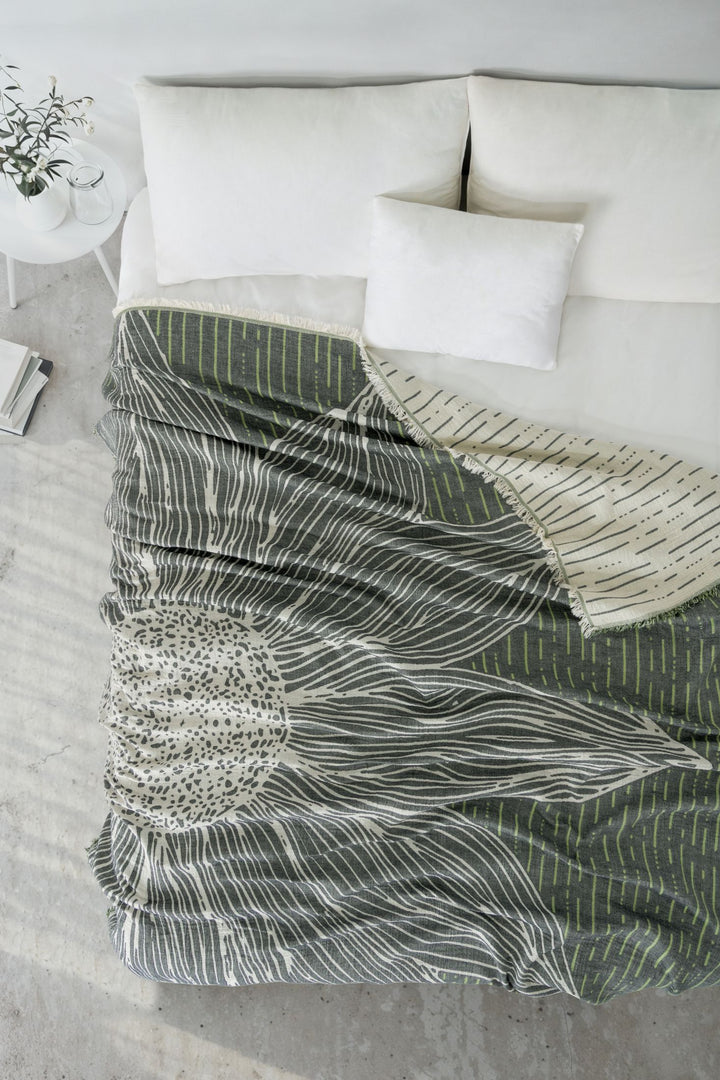 Overhead view of a neatly made bed with white pillows and the Sunflower Bedspread in Green by turkanhome.com, featuring a large abstract floral design in green, cream, and black. A small table with a plant and books is beside the bed.