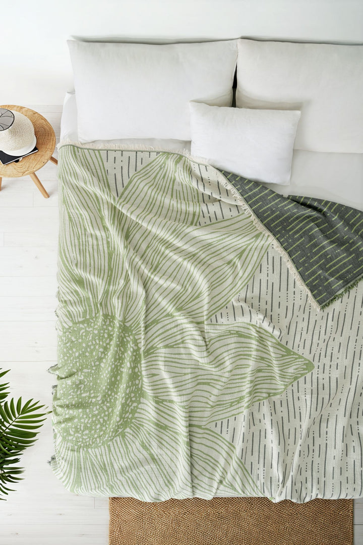 A cozy bed with white pillows and the Sunflower Bedspread in green by turkanhome.com, featuring a bold floral design. Nearby, a small side table holds a hat and book, while a green plant rests on a woven rug beside the bed.