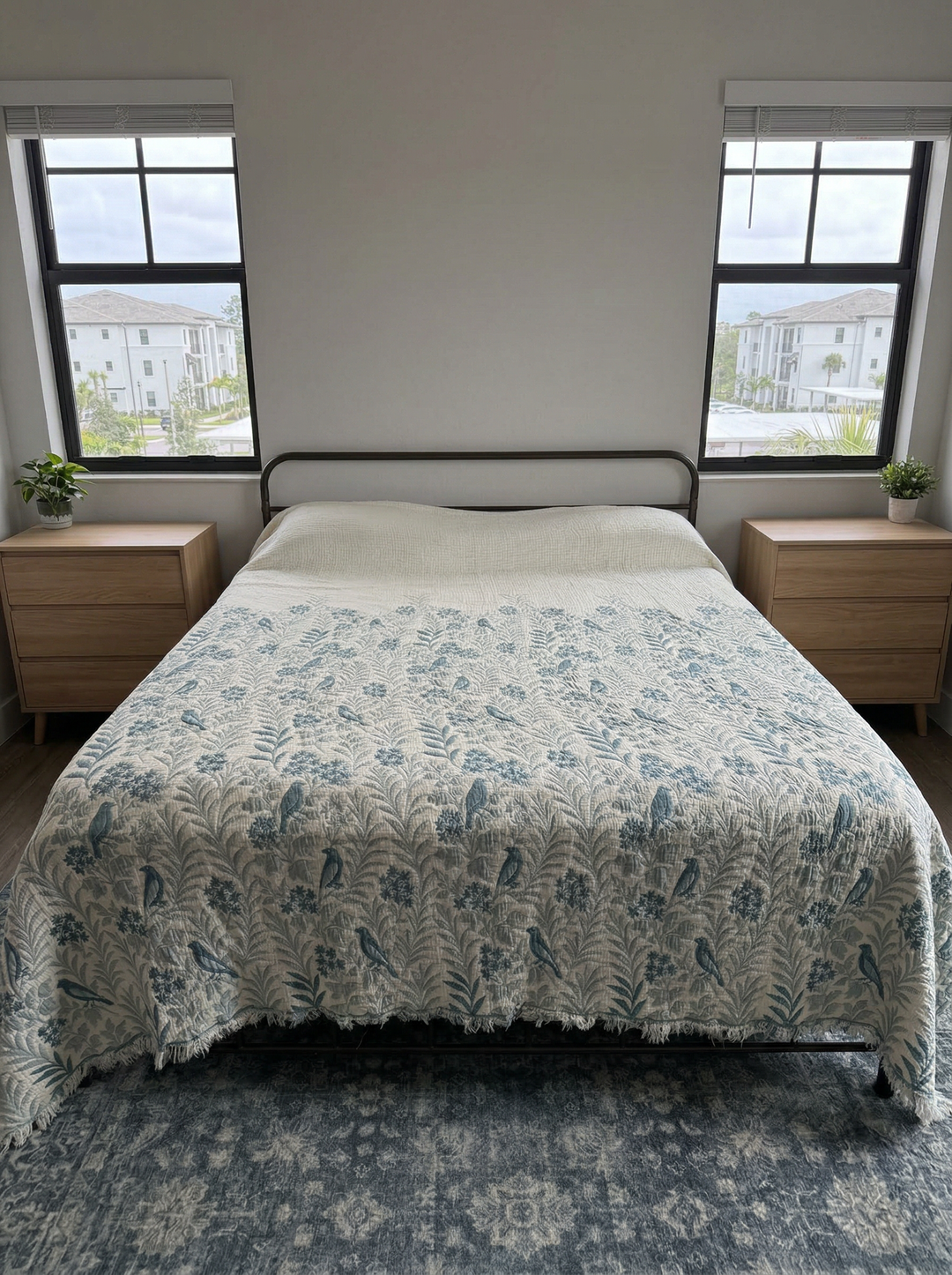A neatly made bed features the turkanhome.com Luxury 7-Layer Cotton Muslin Quilted Blanket in Mint Bird design, nestled between wooden nightstands with potted plants. Two windows behind reveal city apartment buildings and greenery outside.