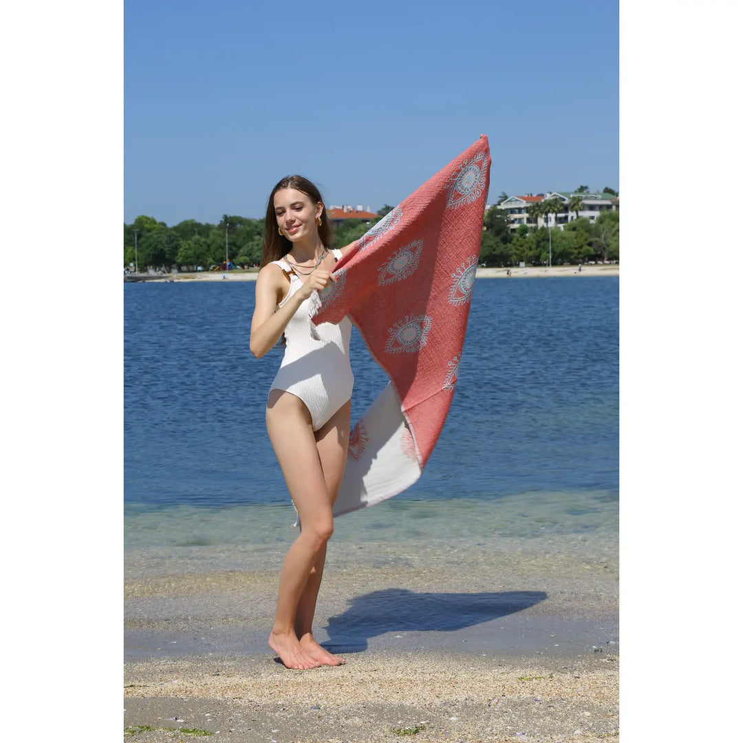 A woman in a white swimsuit stands barefoot on a sandy beach, smiling and holding the Eye Red Sand Resistant Turkish Beach Towel by turkanhome.com. Calm water and green trees can be seen in the background.