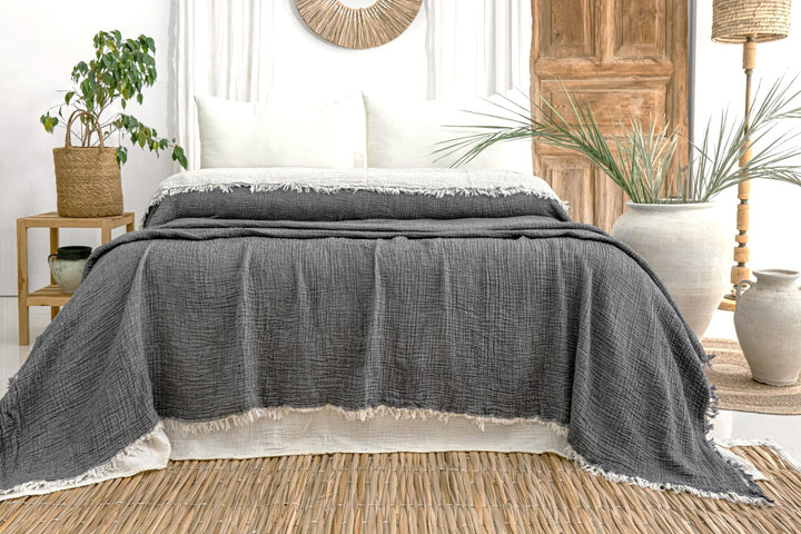 A neatly made bed with white pillows and a turkanhome.com Anthrachite Crinkled Muslin Bed Blanket in a bright, boho-style room with plants, woven decor, and wooden accents.