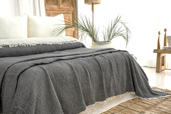 A bed features the Anthrachite Crinkled Muslin Bed Blanket by turkanhome.com—textured dark gray Turkish cotton with white fringe—topped with white pillows, surrounded by wood accents, green plants, a woven rug, and rustic furniture.