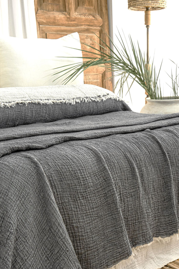 A cozy bed features the Anthrachite Crinkled Muslin Bed Blanket from turkanhome.com, textured in gray and draped over a rustic wooden headboard. A white pillow and tall green leaves complete the natural, relaxed look.