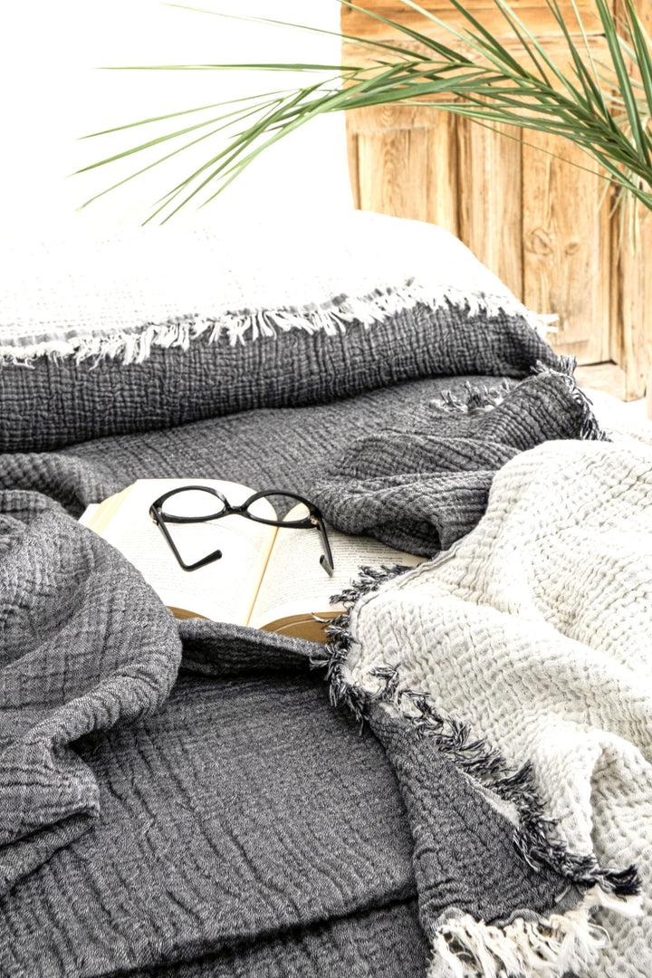 A pair of black eyeglasses rests on an open book atop the Anthrachite Crinkled Muslin Bed Blanket by turkanhome.com, featuring grey and white textures, with a leafy plant and wooden furniture in the bright background.