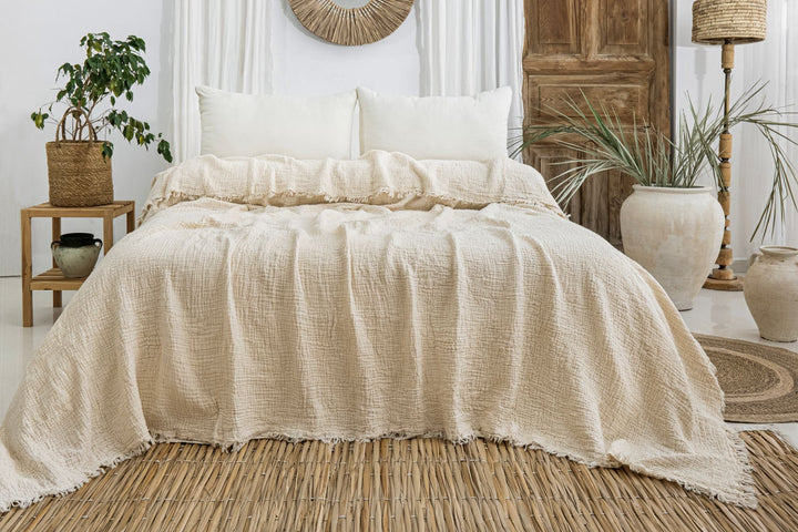 A neatly made bed features the Cream Crinkled Muslin Bed Blanket by turkanhome.com and two white pillows, set in a neutral room with rustic decor—potted plants, a wicker rug, and natural wood accents.