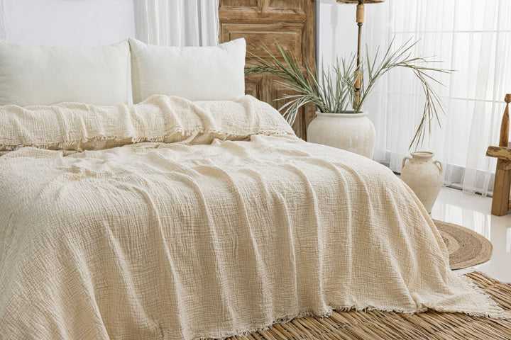 A cozy bed with the Cream Crinkled Muslin Bed Blanket from turkanhome.com, white pillows, and natural accents like a woven rug, potted plants, and pottery brighten this sunlit bedroom.