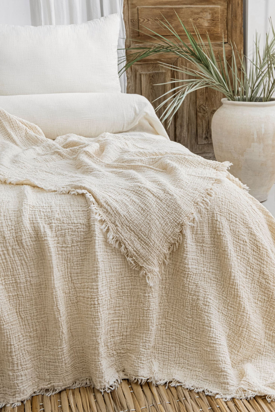 The Cream Crinkled Muslin Bed Blanket from turkanhome.com, in soft cream Turkish cotton with fringed edges, rests on white bedding. Nearby is a large ceramic vase with green leaves and a wooden door in the background.