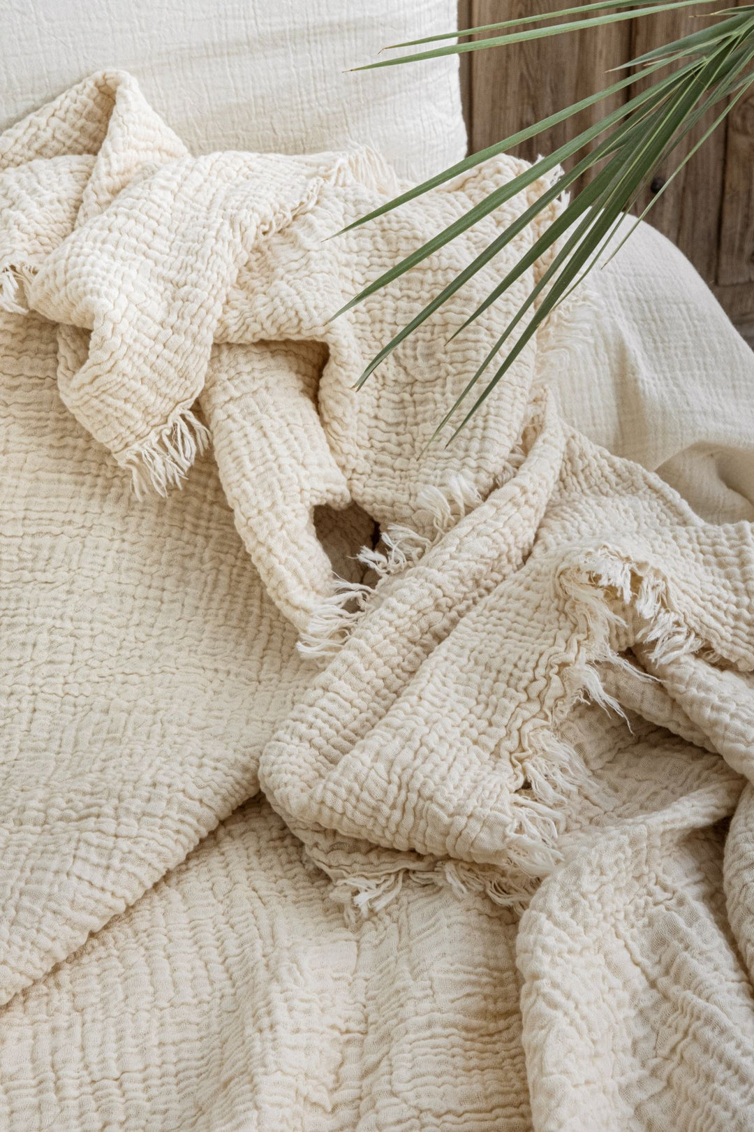 The Cream Crinkled Muslin Bed Blanket from turkanhome.com, in a soft cream hue with fringe edges, is draped over a bed while a green palm leaf peeks in at the upper right corner.