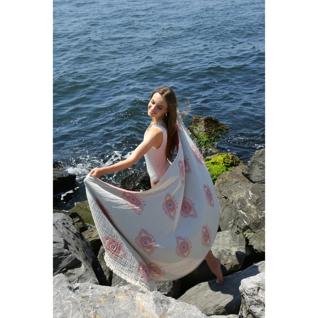 A woman in a white swimsuit smiles on a rocky shoreline, holding the Eye Red Sand Resistant Turkish Beach Towel by turkanhome.com as it billows in the ocean breeze with waves and mossy rocks creating a stunning coastal backdrop.