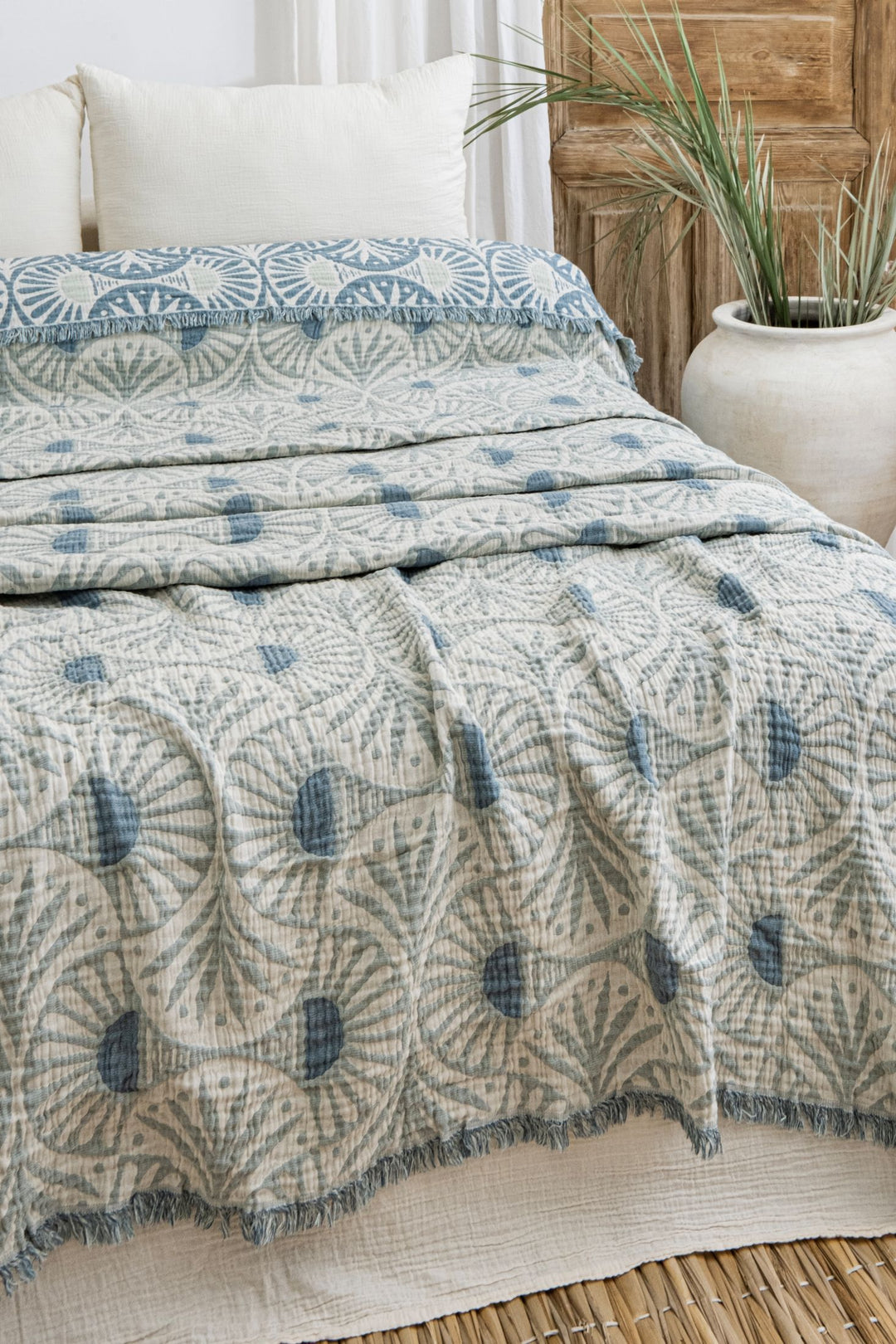 A bed features the Gingko Muslin Bed Blanket in teal from turkanhome.com, with matching pillows, next to a large white planter holding a green plant, near a wooden door and sheer white curtains in the background.