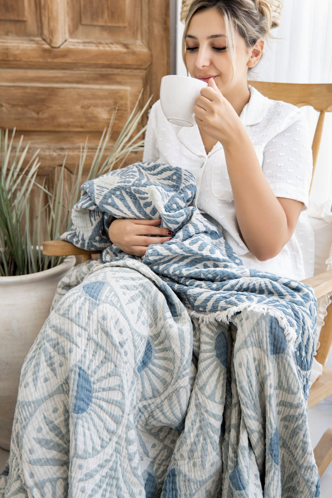A woman relaxes by a wooden door, wrapped in the Gingko Muslin Bed Blanket – Teal from turkanhome.com. She smiles with her eyes closed, holding a white mug, seated on a wooden chair near a large potted plant, and wearing a white top.