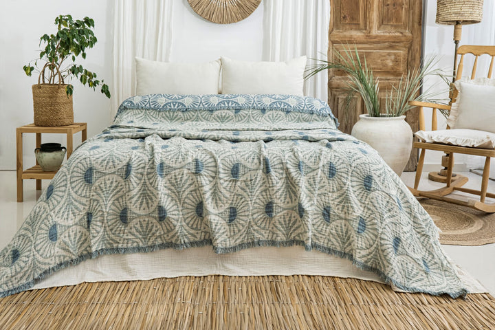 A cozy bed with white pillows and the Gingko Muslin Bed Blanket – Teal (King-Oversized Queen) from turkanhome.com creates a fresh, bohemian vibe alongside plants, a wicker chair, and natural wood décor in a light, airy room.