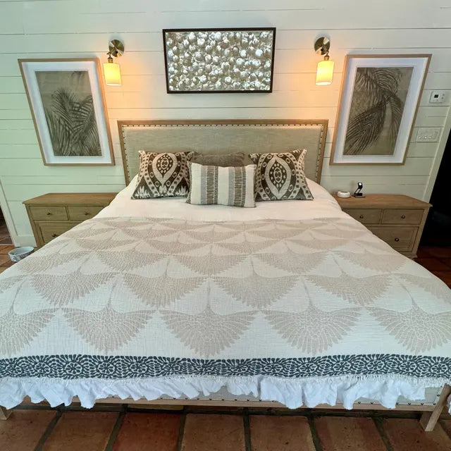 A neatly made bed with patterned pillows and the Crane Muslin Bed Blanket in Ivory from turkanhome.com sits between two wooden nightstands, beneath three framed artworks and two wall-mounted lamps on a white paneled wall.