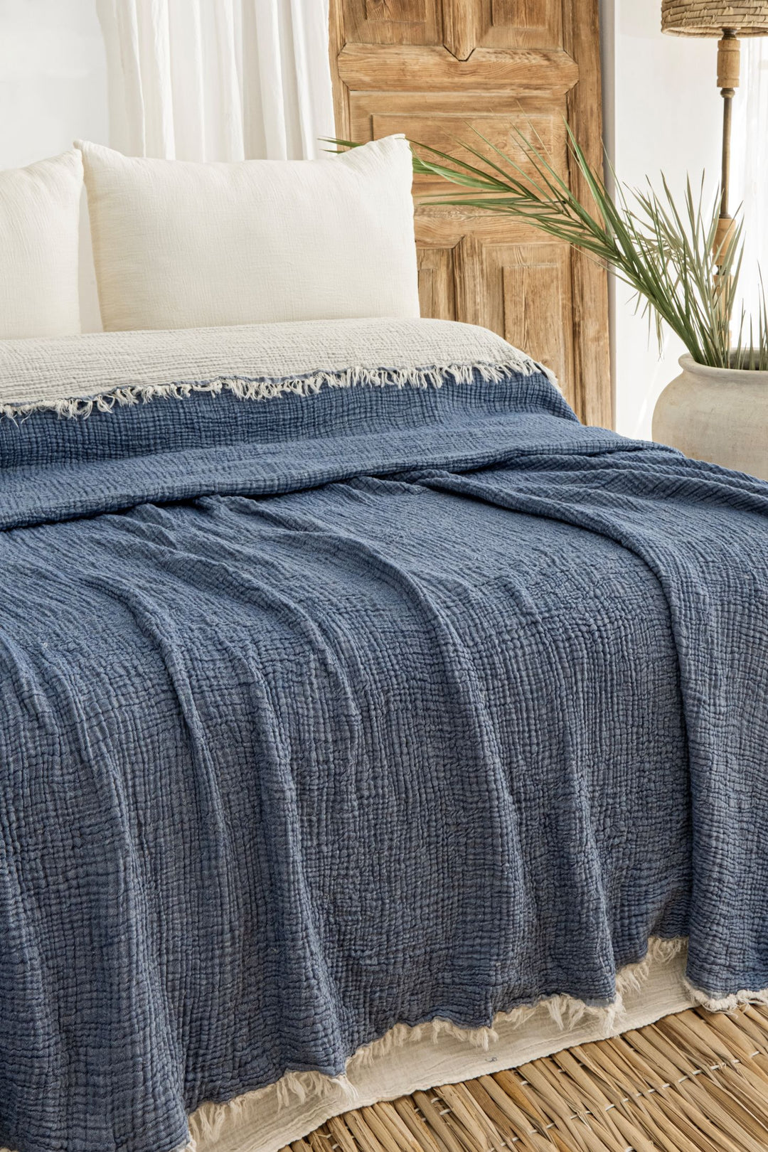 A neatly made bed features cream pillows and the Indigo Crinkled Muslin Bed Blanket from turkanhome.com, layered with a textured blue throw. The room includes a wooden door, sheer white curtains, a large potted plant, and a woven rug.