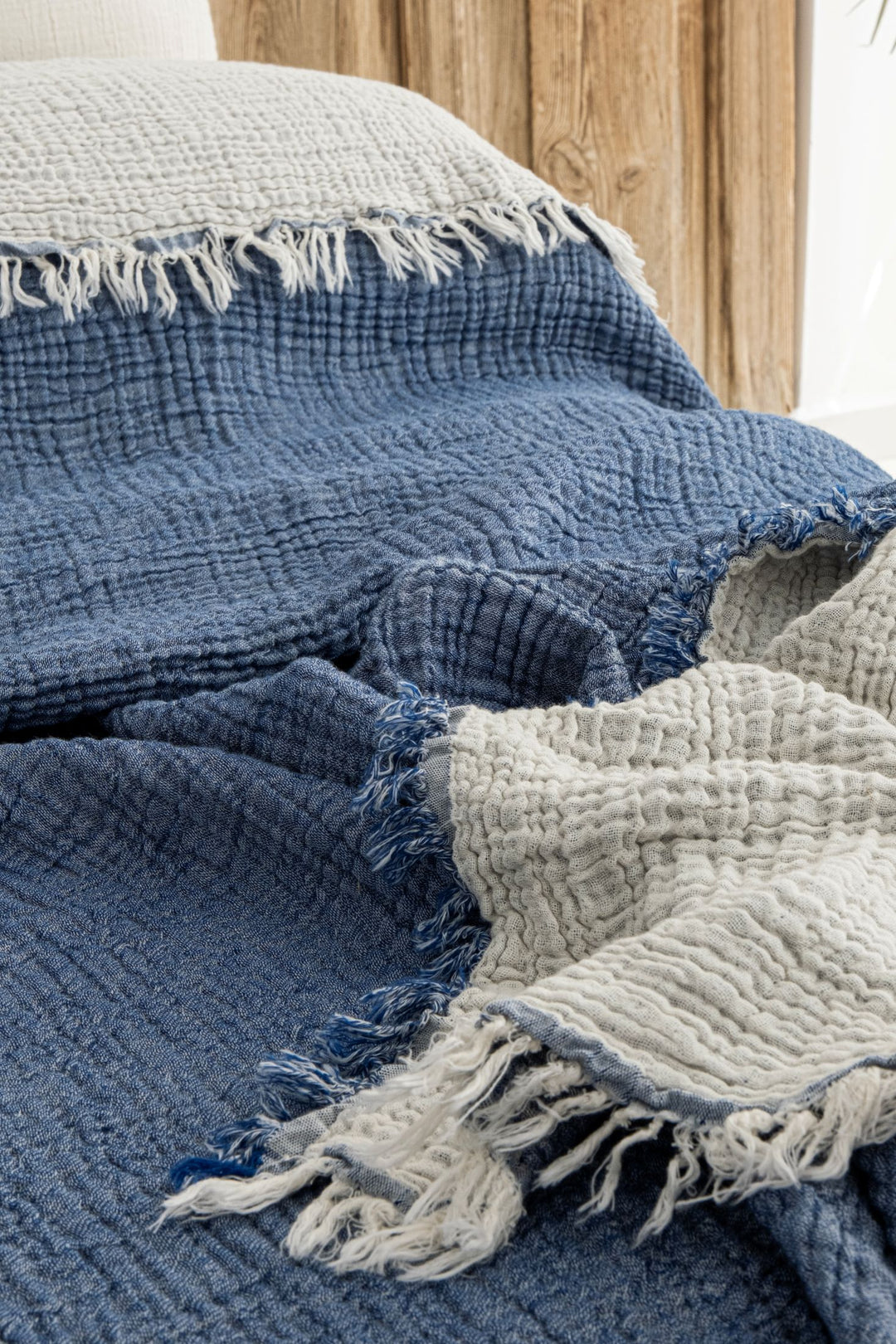 The Indigo Crinkled Muslin Bed Blanket by turkanhome.com, featuring blue and beige sections, a textured crinkled pattern, and fringed edges, is draped over a bed with a wooden headboard and part of a white pillow in the background.