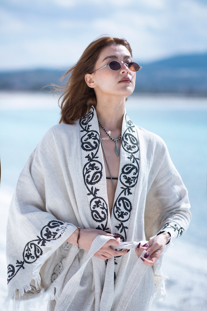 A woman in sunglasses wears the Laodikya - Lightweight Yoga Kimono from turkanhome.com by a bright blue sea, her hair blowing in the wind. Blurred mountains and sky appear in the softly sunlit background.
