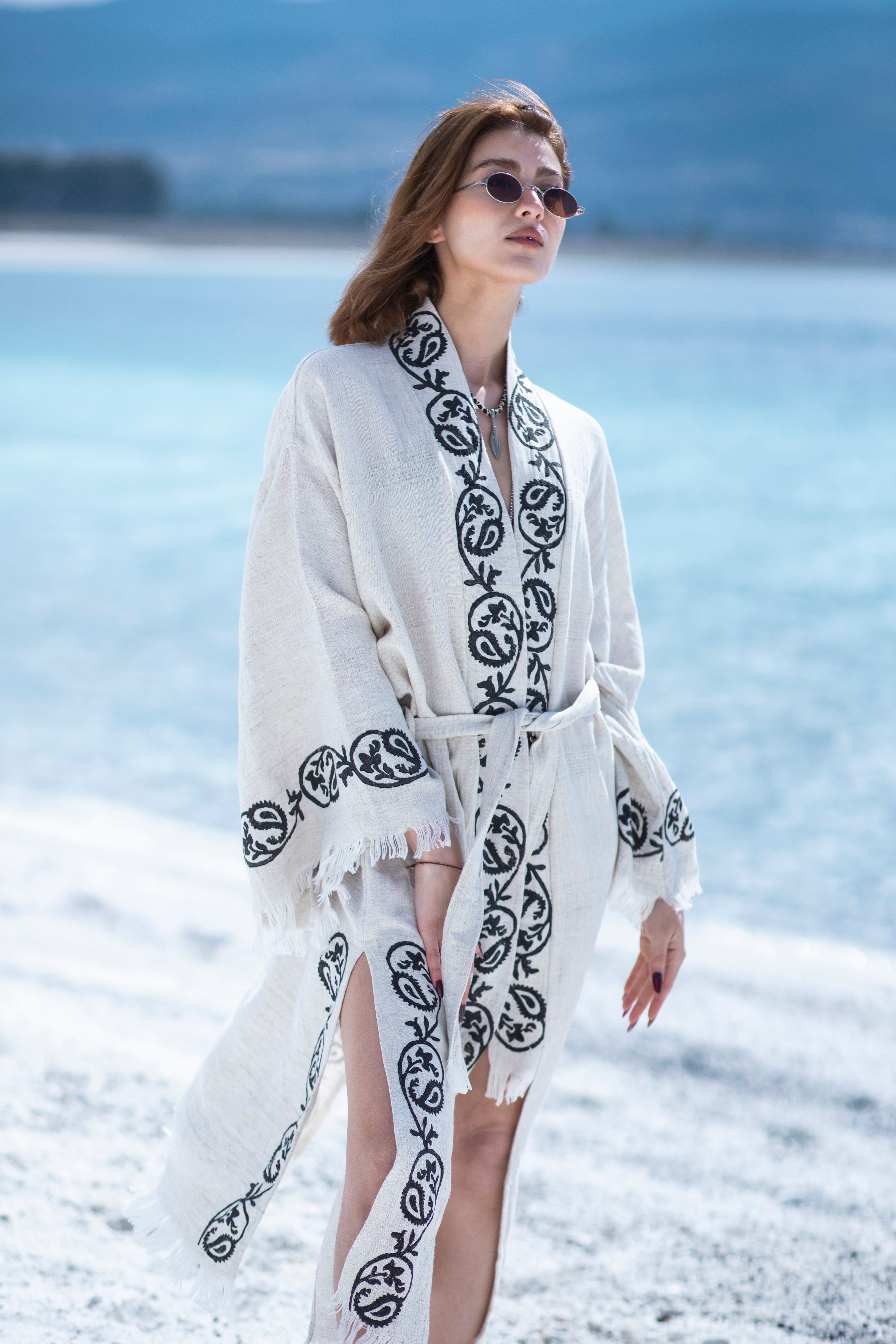 A woman in the Laodikya - Lightweight Yoga Kimono from turkanhome.com, styled with round sunglasses, stands on a beach with bright blue water and distant mountains in the background.