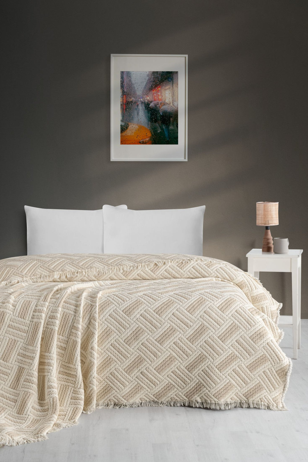 A neatly made bed with a cream Comfort Muslin Quilt from turkanhome.com and white pillows stands against a dark gray wall. A framed abstract painting hangs above, while a small white nightstand with a lamp sits beside it.