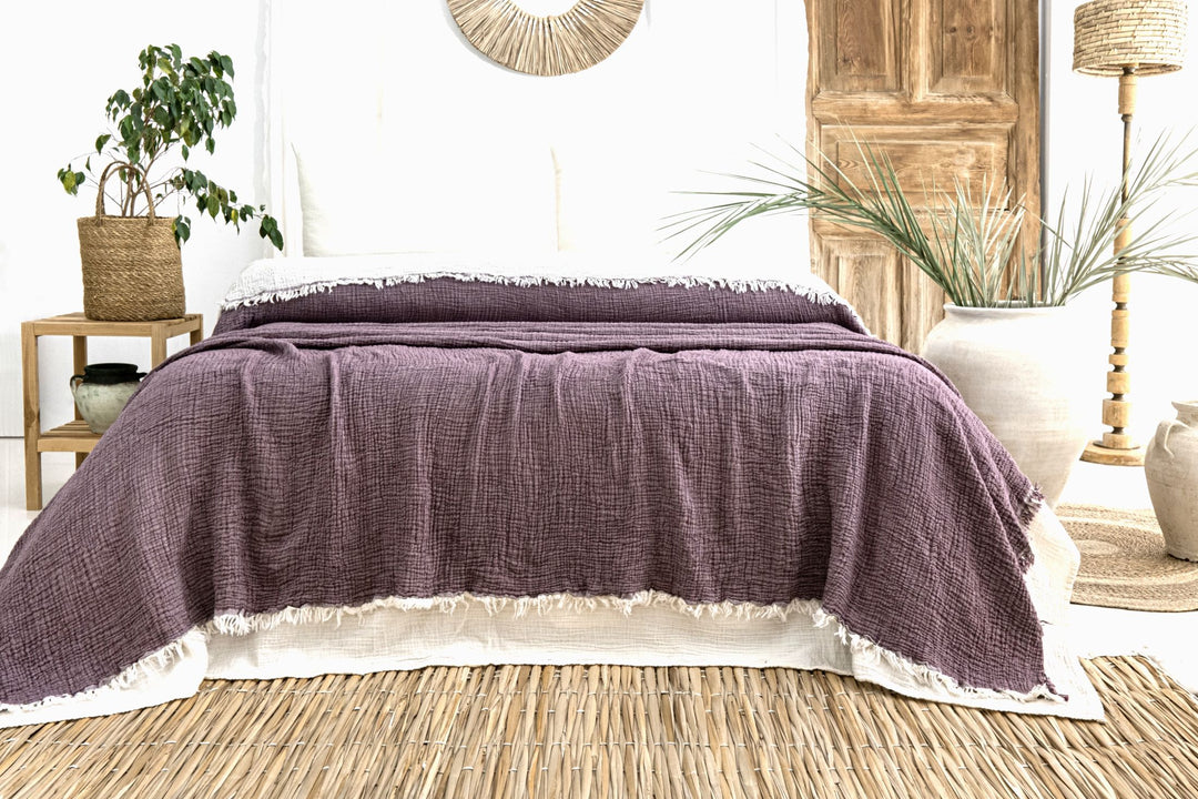 A neatly made bed with the Purple Crinkled Muslin Bed Blanket from turkanhome.com stands in a bright, bohemian-style room featuring wooden decor, plants, and natural woven accents.