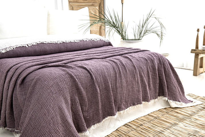 A bed features the Purple Crinkled Muslin Bed Blanket by turkanhome.com with white fringed edges, white pillows, and a woven rug. Wooden furniture and a potted plant in the background create a cozy, natural vibe.