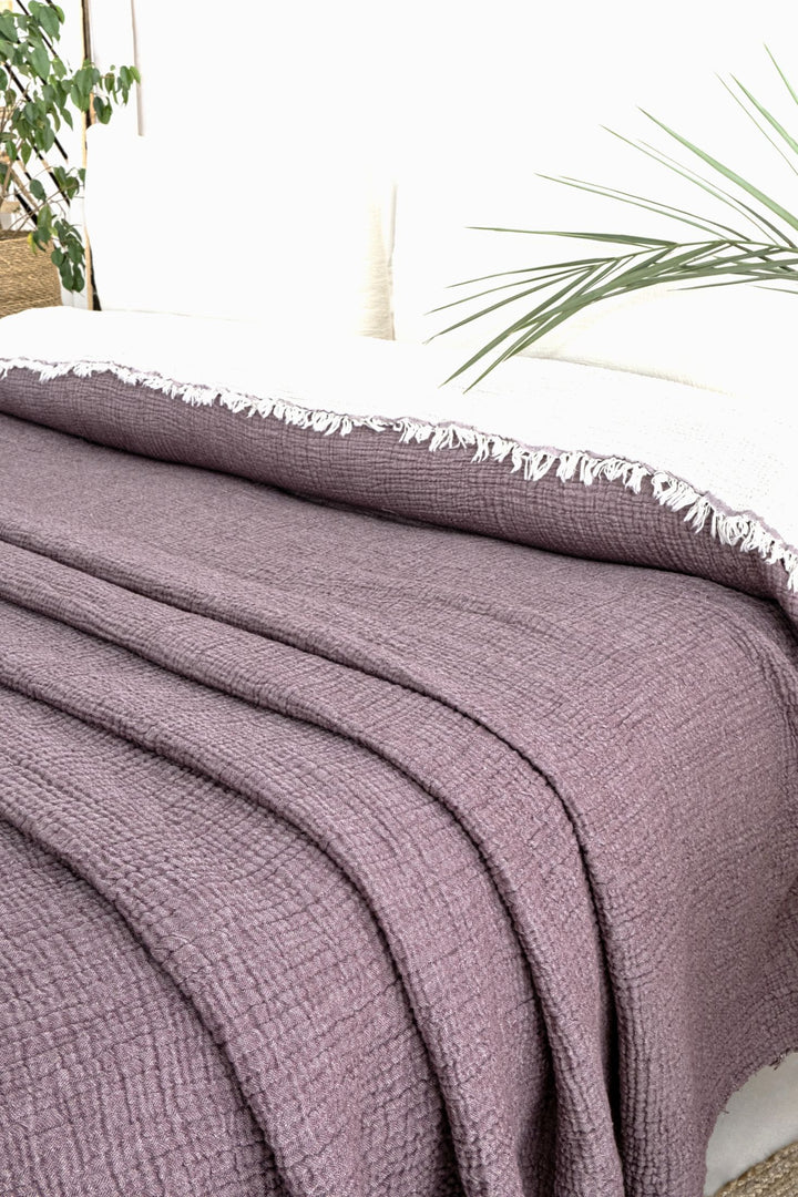 A neatly made bed features the turkanhome.com Purple Crinkled Muslin Bed Blanket with a white fringed throw at the head. White pillows and a green plant leaf in the background add to the cozy, fresh vibe.