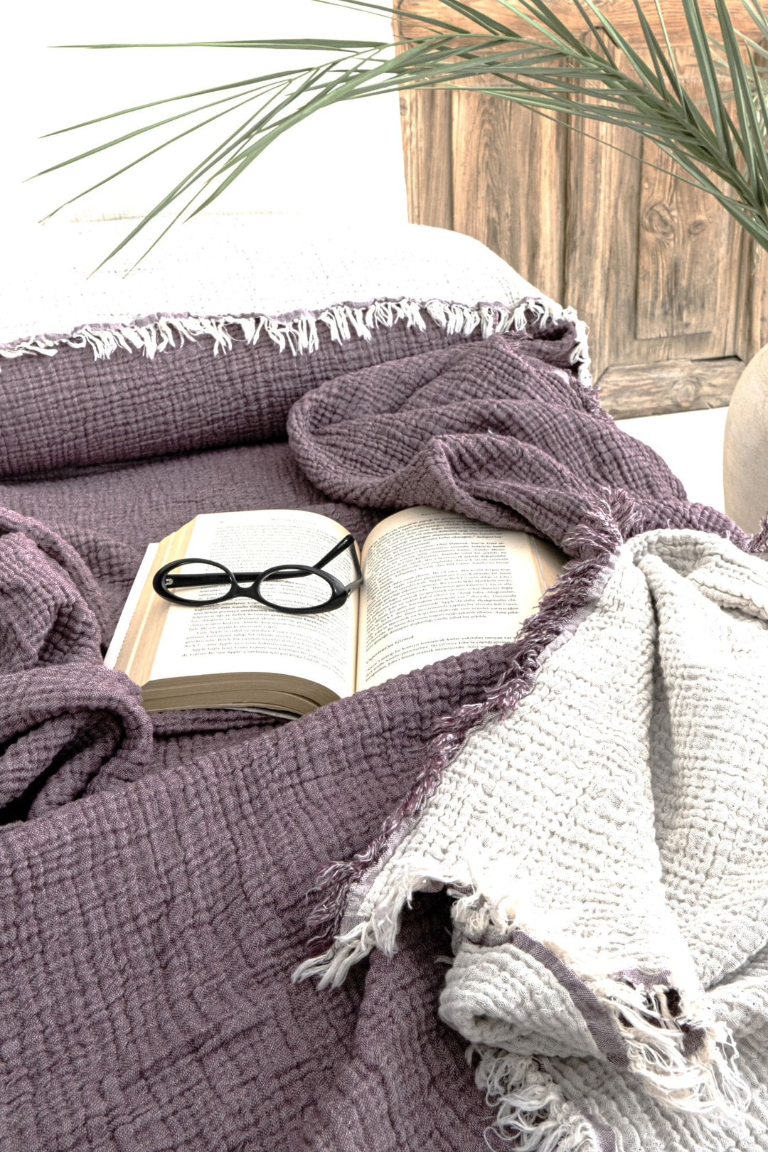 A pair of black eyeglasses sits on an open book atop the Purple Crinkled Muslin Bed Blanket by turkanhome.com, with a green plant and wooden surface in the background.