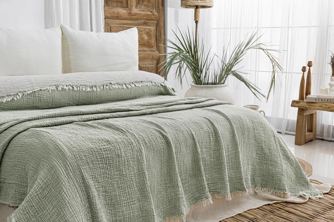 A bright, airy room with wooden decor and potted plants features a bed dressed in light green bedding and white pillows, topped with the Sage Crinkled Muslin Bed Blanket from turkanhome.com, basking in sunlight through sheer curtains.