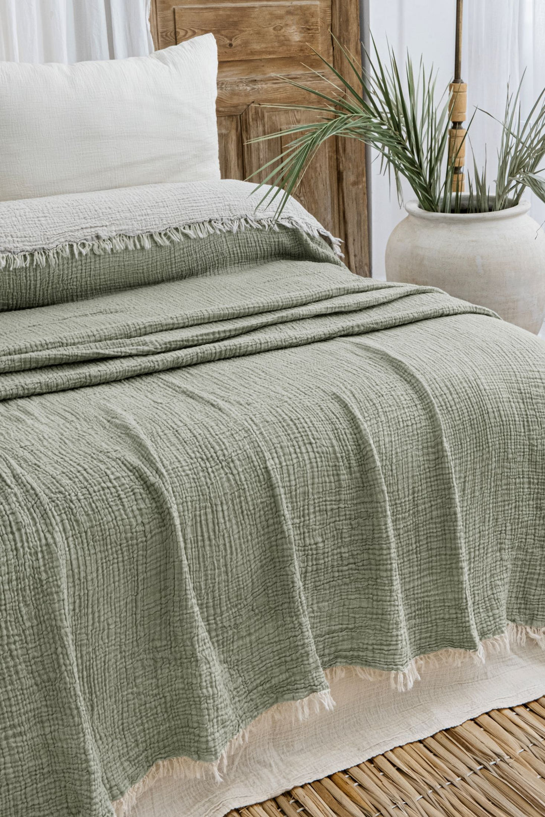 A bed with white pillows and the Sage Crinkled Muslin Bed Blanket from turkanhome.com, featuring fringed edges, rests on a woven rug. Behind the bed is a rustic wooden door and a large white vase with green plants.