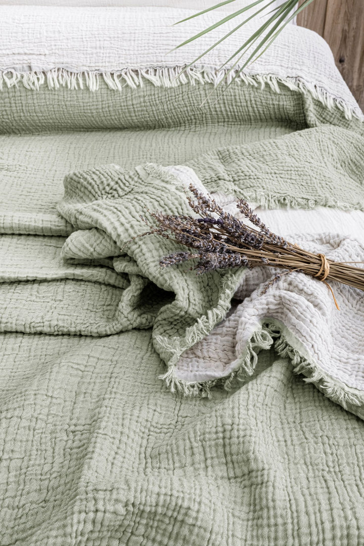 The Sage Crinkled Muslin Bed Blanket by turkanhome.com, featuring soft green and white Turkish cotton with fringed edges, is displayed on a bed with a dried lavender bouquet and a green leaf in the corner.
