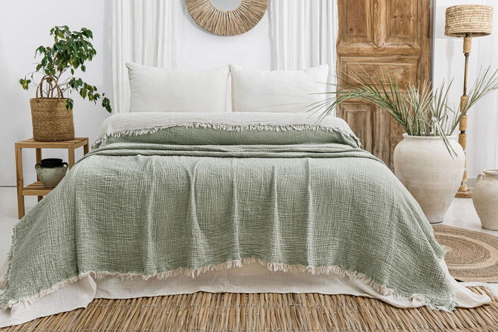 A cozy bedroom features a neatly made bed with white pillows and the Sage Crinkled Muslin Bed Blanket from turkanhome.com, surrounded by potted plants, wicker accents, a wooden door, and neutral-toned natural decor.