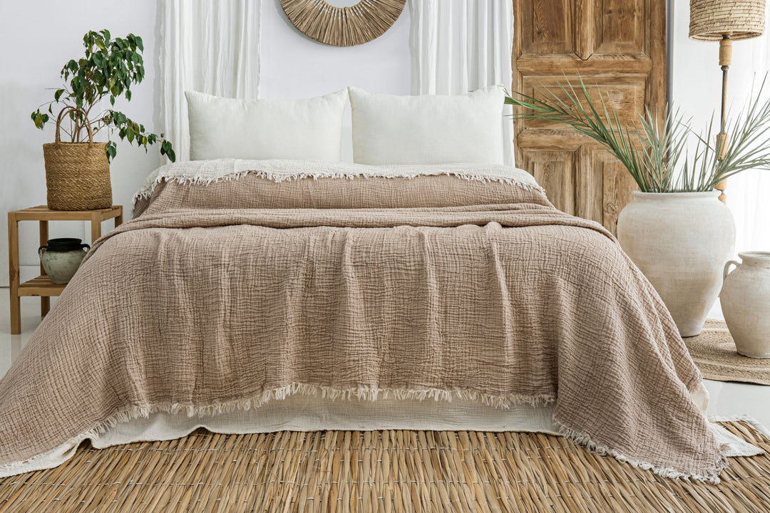 A cozy bed featuring turkanhome.com’s Taupe Crinkled Muslin Bed Blanket with cream pillows, set in a rustic room with wooden accents, potted plants, a woven rug, and neutral decor.