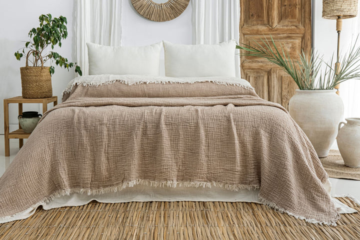 A cozy bed featuring turkanhome.com’s Taupe Crinkled Muslin Bed Blanket with cream pillows, set in a rustic room with wooden accents, potted plants, a woven rug, and neutral decor.
