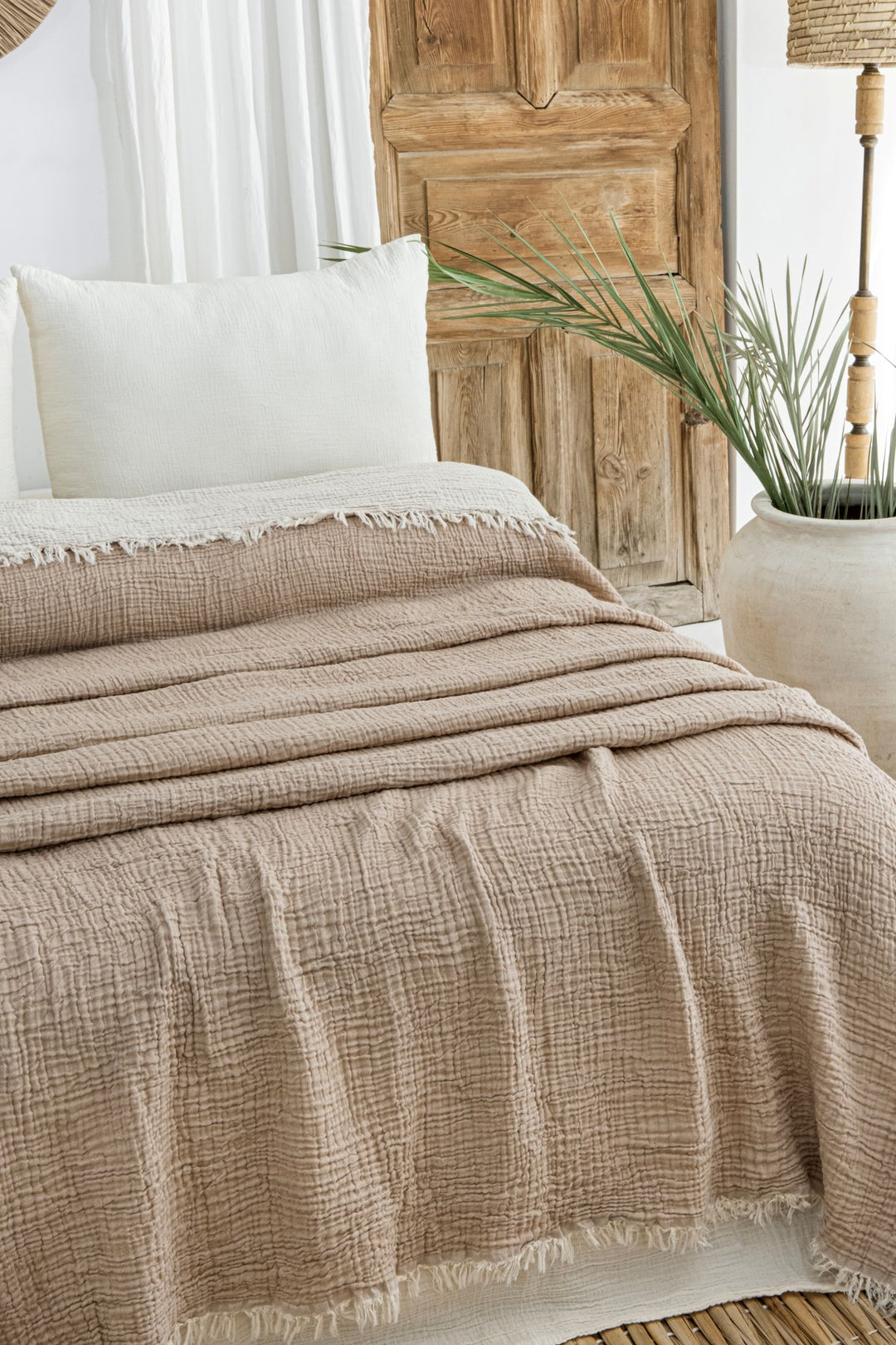 A neatly made bed with a Taupe Crinkled Muslin Bed Blanket from turkanhome.com and a white pillow sits beside a large white vase with green palm leaves, all in front of a rustic wooden door.