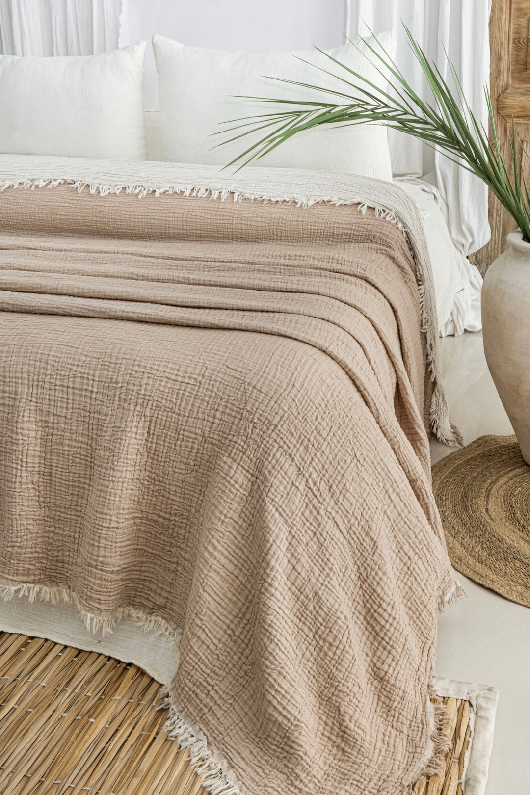 A neatly made bed features the Taupe Crinkled Muslin Bed Blanket by turkanhome.com with white pillows, beside a large vase with a green palm leaf, all set atop a natural woven rug in a bright, airy room.
